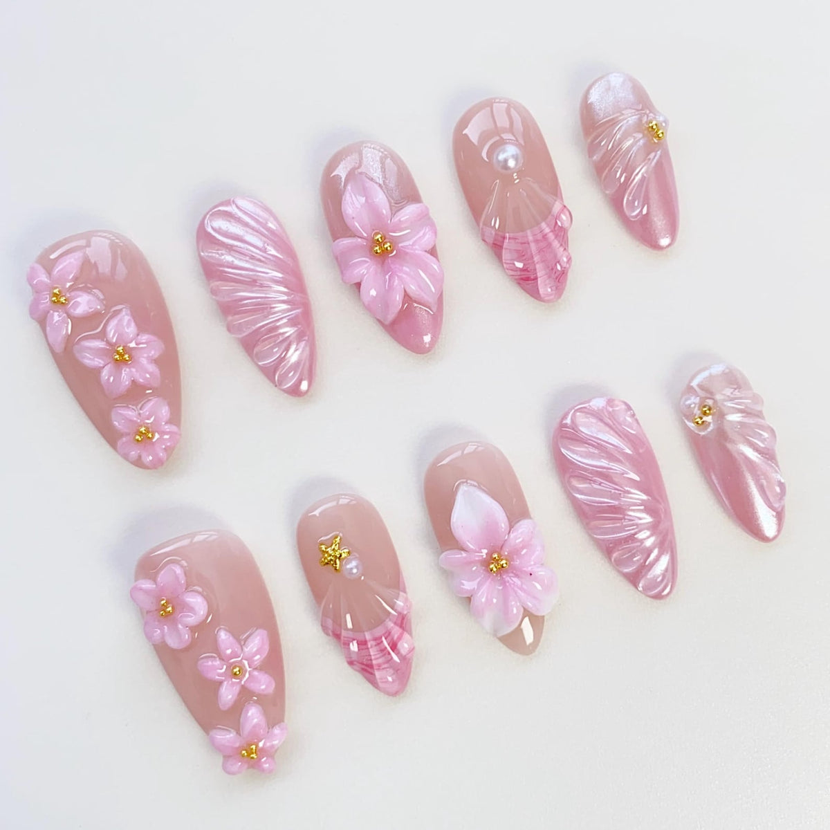 A set of ten almond-shaped artificial nails. Each nail is painted in a light pink hue and adorned with 3D floral designs, pearl accents, and textured patterns. The nails are arranged on a white surface.