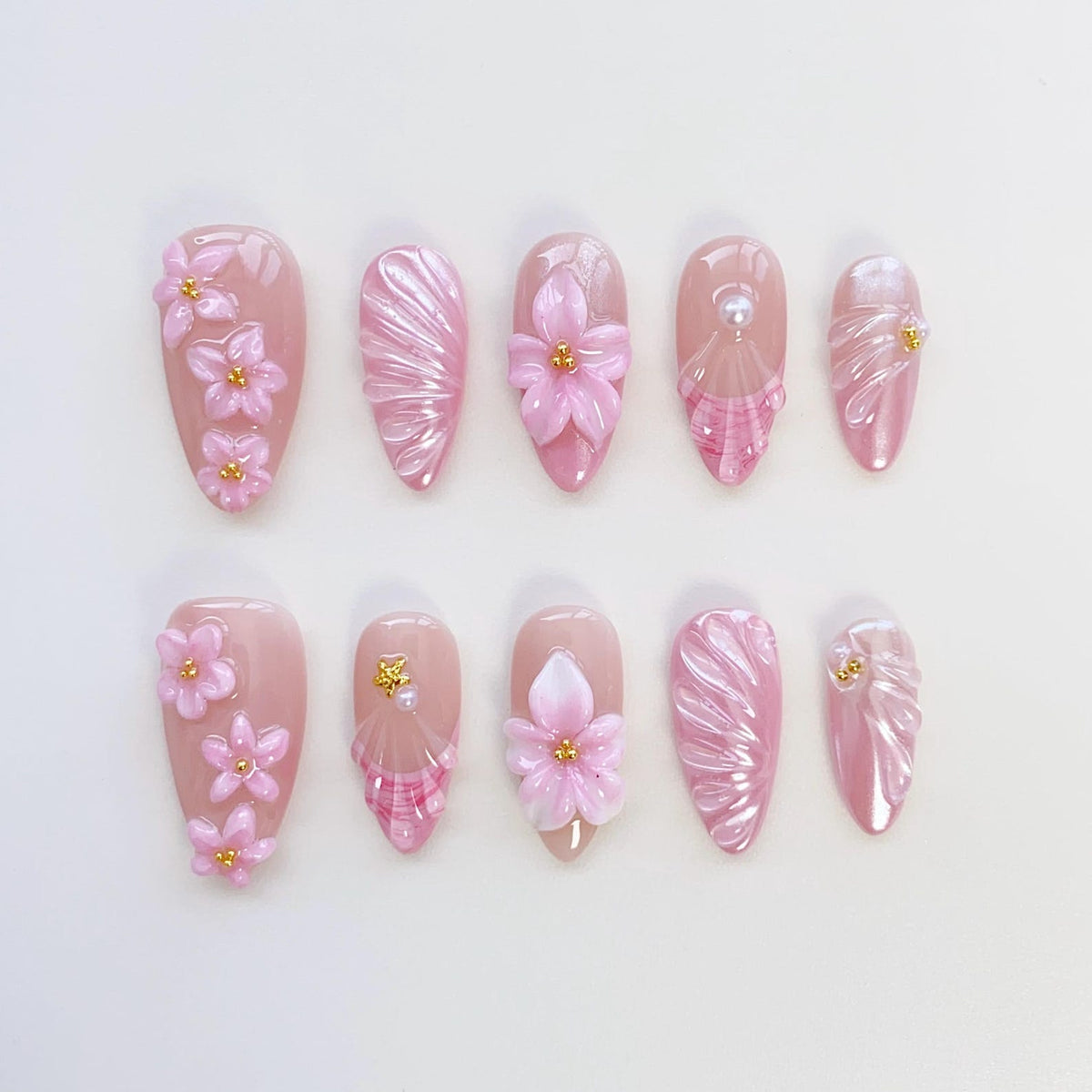 A set of ten artificial nails in various designs. The nails are primarily pink, with some featuring 3D pink flowers, pearl accents, and textured patterns. Each nail has a glossy finish.