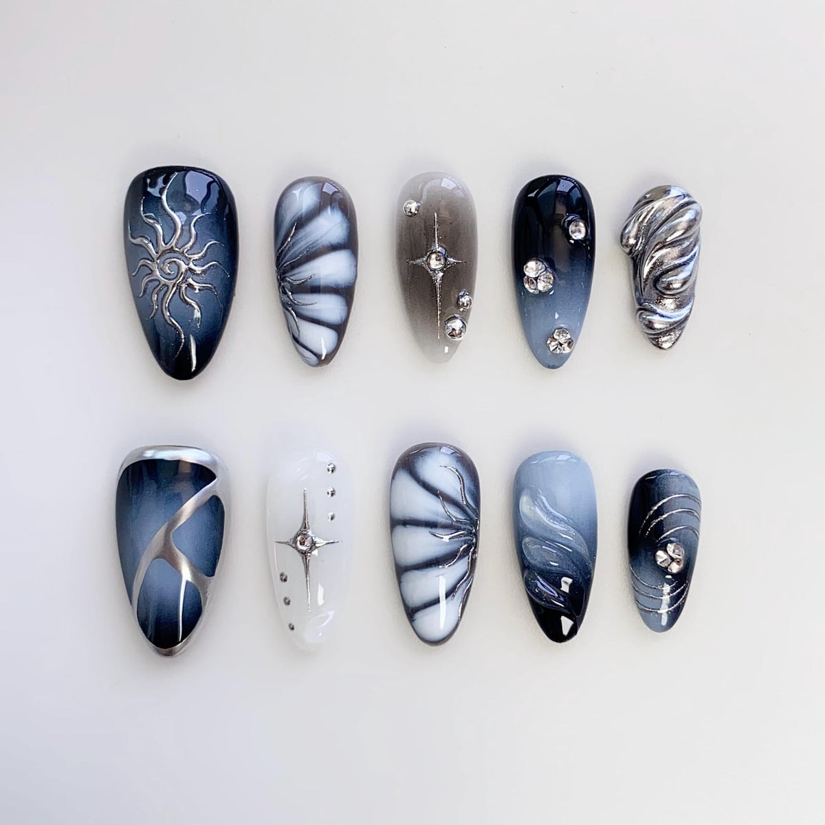 A set of ten artificial nails in various designs. The nails feature a range of colors, including black, white, and gray, with decorative elements such as silver swirls, starbursts, and rhinestones. The nails are oval-shaped.