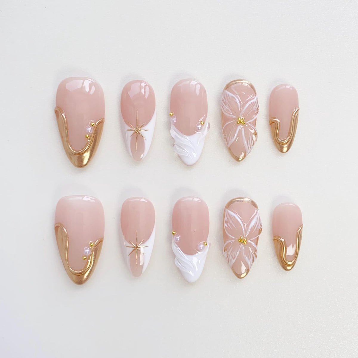 A set of ten almond-shaped false nails. The nails feature a pale pink base with gold accents, white floral designs, and small pearl embellishments. Some nails have a gold border.