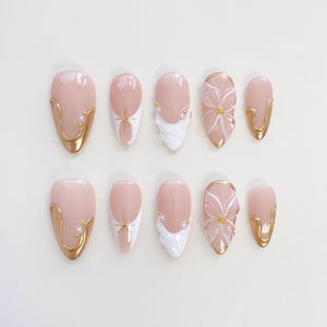 A set of ten almond-shaped false nails. The nails feature a pale pink base with gold accents, white floral designs, and small pearl embellishments. Some nails have a gold border.