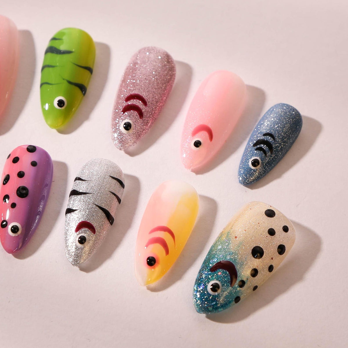 A collection of artificial nails with various designs. The nails feature fish-themed art, including green, pink, and blue glitter, with details like eyes and scales. Some nails have a solid color, whi