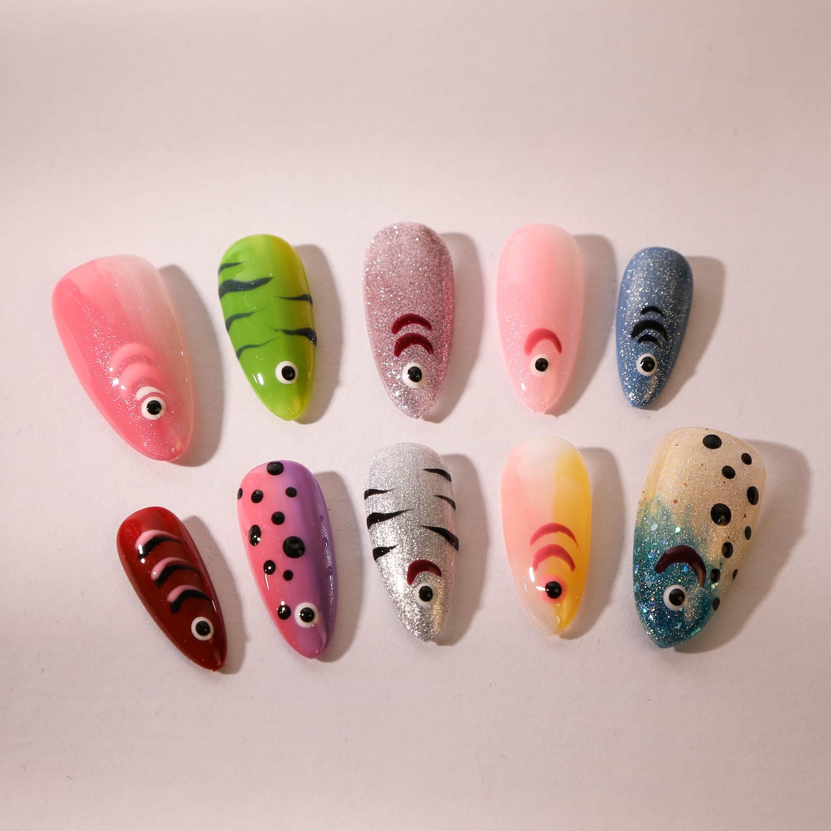 A collection of ten artificial nails, each painted with a unique fish-inspired design. The nails feature a variety of colors, including pink, green, silver, and blue, with details like eyes and scales