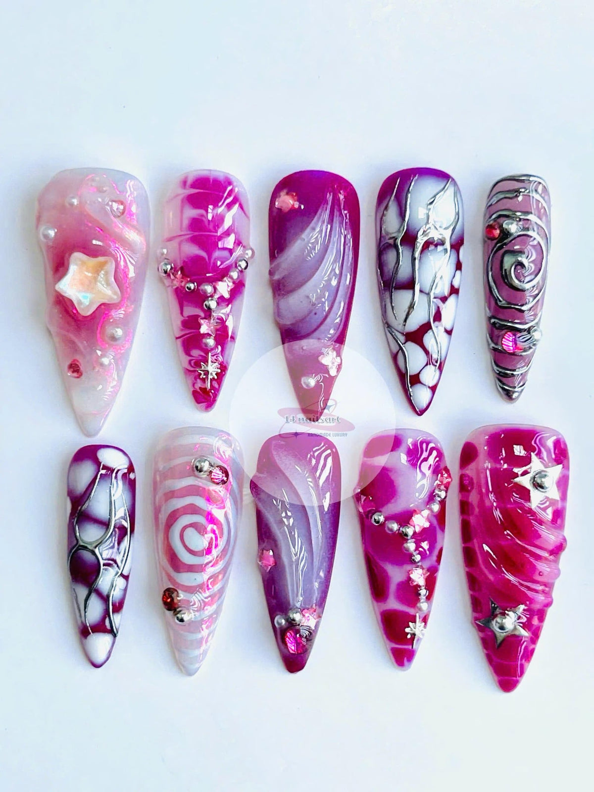 A collection of ten long, pointed artificial nails with various designs. The nails feature shades of pink, purple, and white, adorned with glitter, beads, and star-shaped embellishments. Each nail sho