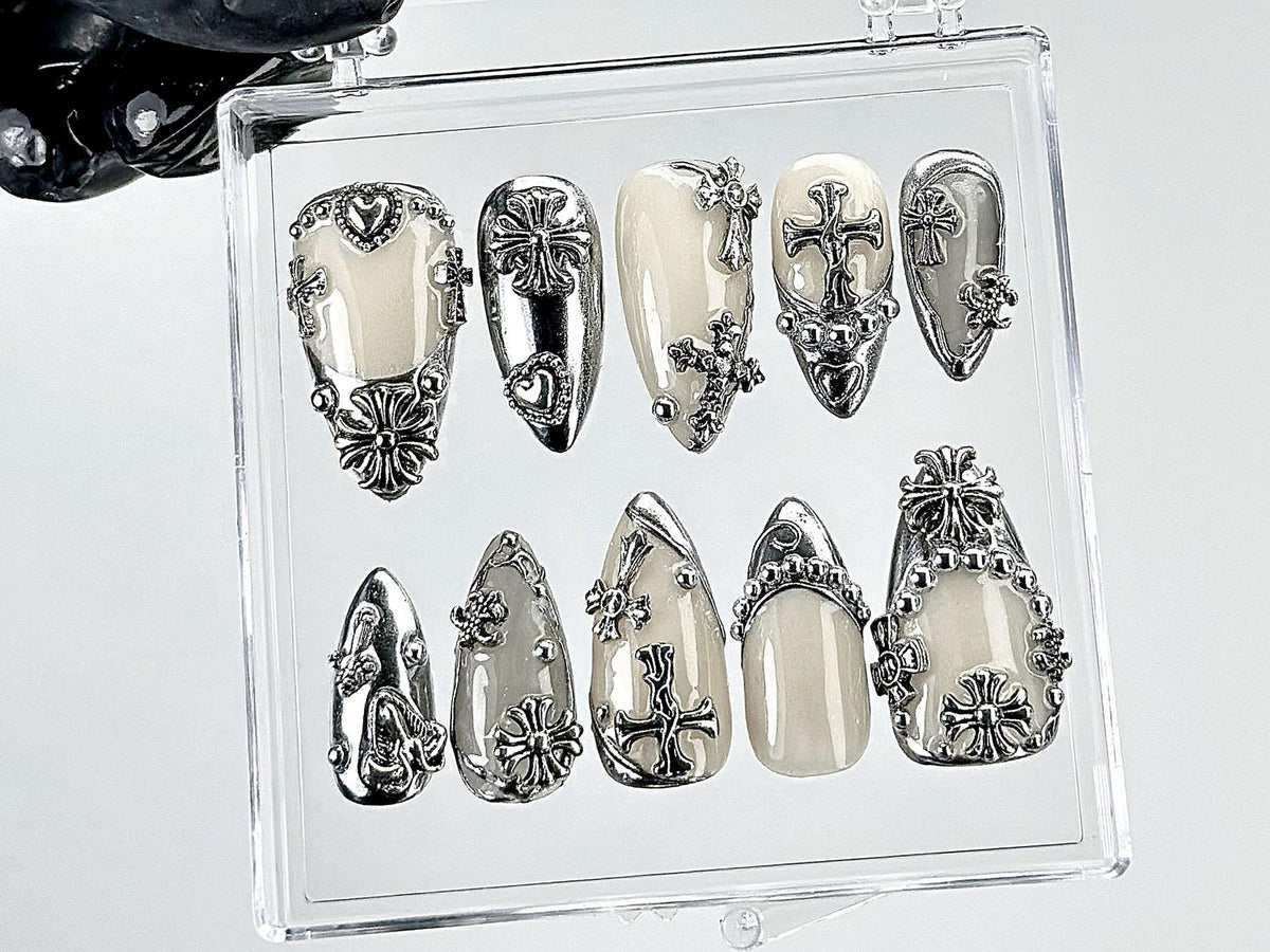 A set of ten artificial nails in a clear plastic case. The nails are various shapes and feature a cream-colored base with silver-colored cross and floral embellishments. Some nails have heart-shaped a