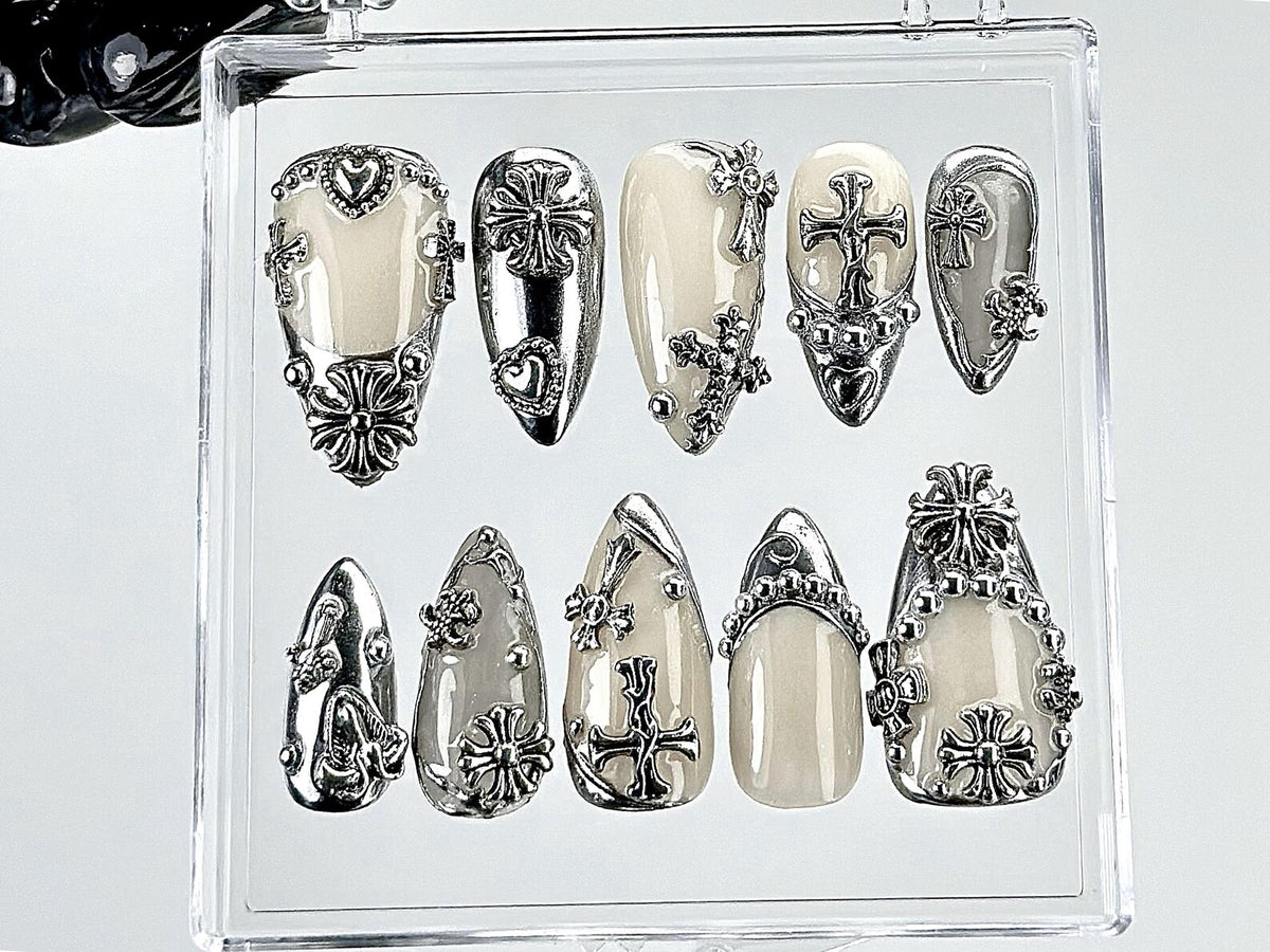 A set of ten artificial nails in a clear plastic case. The nails are a light cream color with silver-colored embellishments. The embellishments include crosses, hearts, and floral designs. The nails a