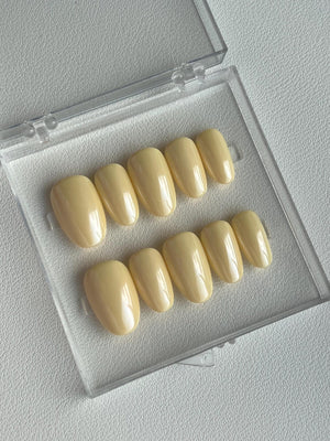 A set of ten pale yellow press-on nails in a clear plastic case. The nails are oval-shaped with a glossy finish. The case is open, revealing the full set of nails.