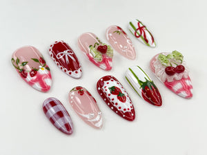 A collection of almond-shaped press-on nails with various designs. The nails feature strawberry, bow, and gingham patterns in red, pink, and white. Some nails have 3D embellishments, including strawbe