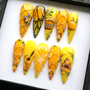 A set of ten yellow acrylic nails with various designs. The designs include a bee, butterfly, honey pot, honeycomb, and a guitar. The nails are decorated with gold accents and floral details.