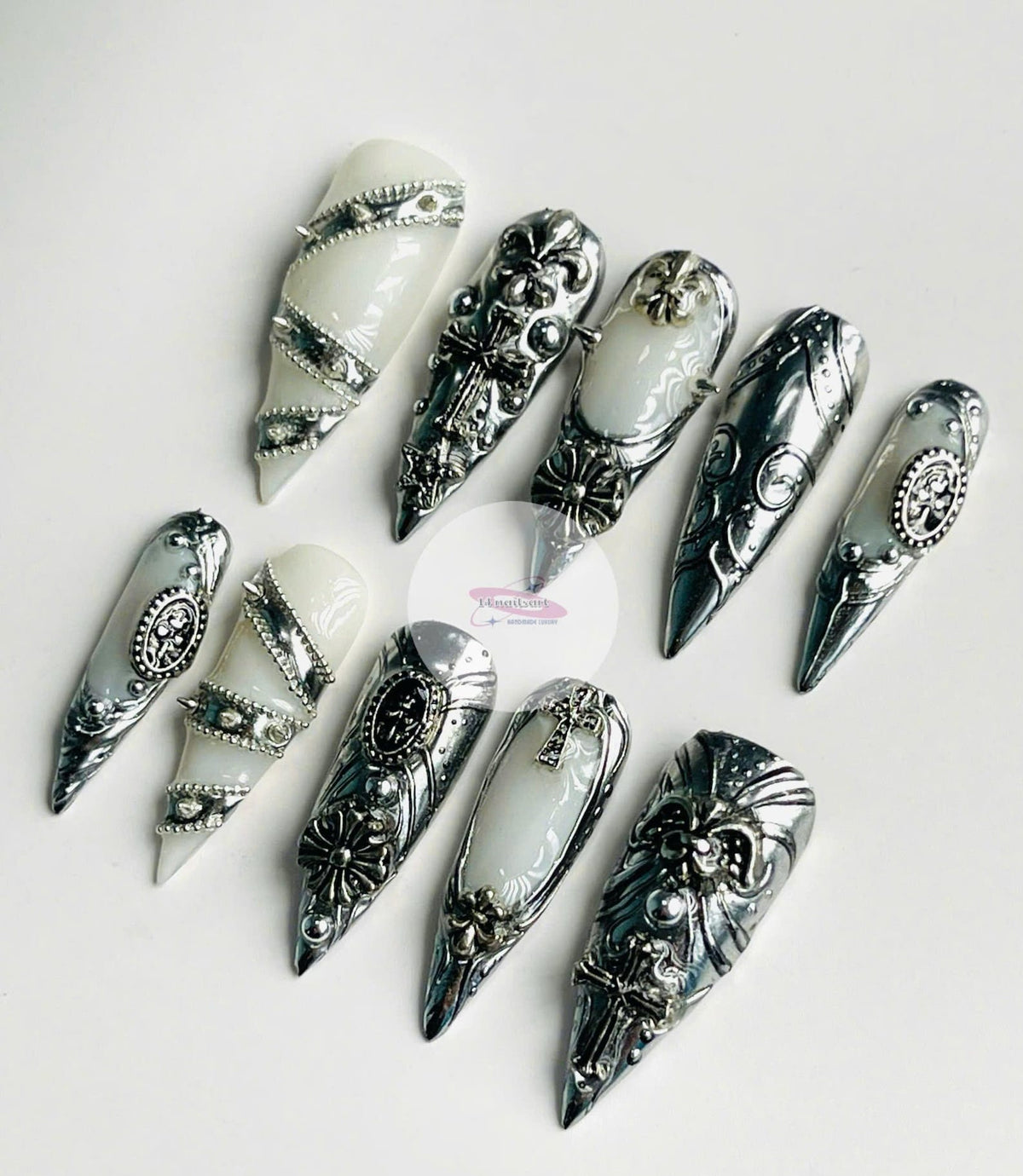 An assortment of silver and white nail art designs. The nail art features ornate metal designs, including crosses, fleur-de-lis, and studded accents. The nails are displayed on a white surface, showca