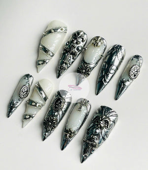 An assortment of silver and white nail art designs. The nail art features ornate metal designs, including crosses, fleur-de-lis, and studded accents. The nails are displayed on a white surface, showca
