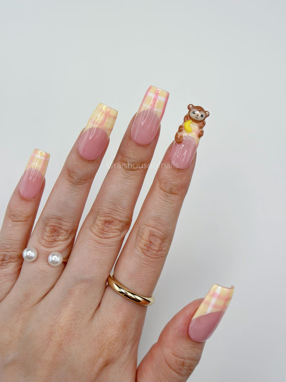 A hand with long, square-shaped artificial nails. The nails feature a pink base with a yellow and white plaid design on the tips. One nail has a small brown monkey figurine. The hand also has a gold r