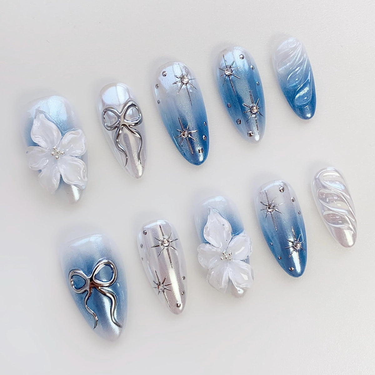 A set of ten artificial nails in various designs. Some nails feature a gradient of blue and white, while others have silver bows, starburst accents, or white floral embellishments. The nails have an oval shape and a glossy finish.