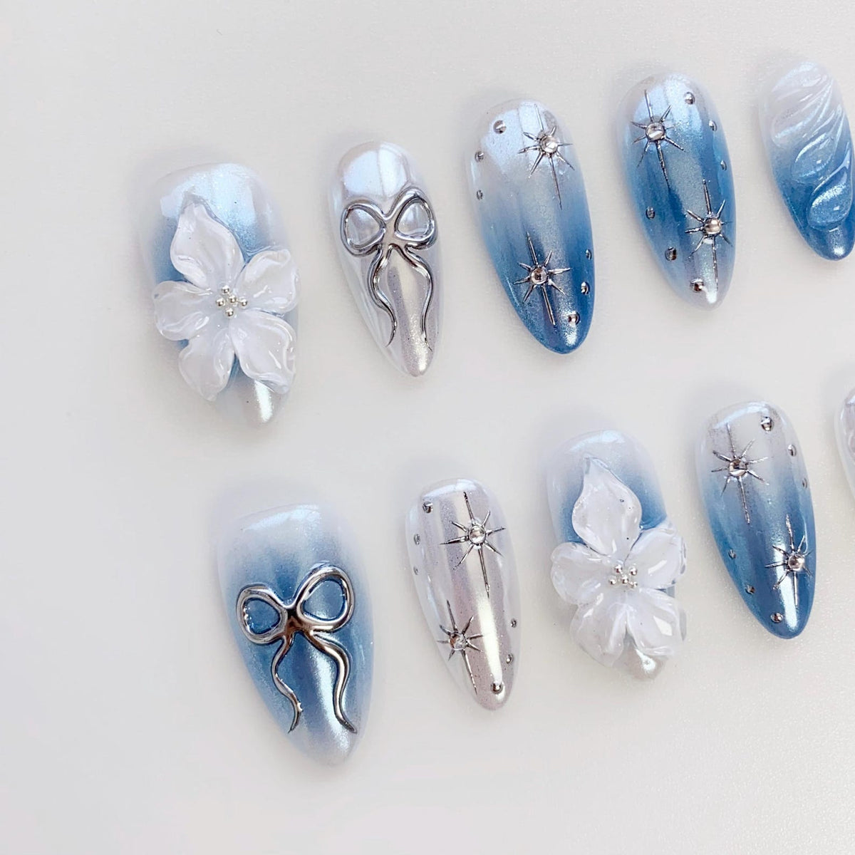 A set of artificial nails with a light blue and white color scheme. Some nails feature 3D white flowers, silver bows, and starburst designs. The nails have an oval shape and a glossy finish, perfect for a special occasion.