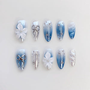A set of ten artificial nails with a light blue and white color scheme. Designs include 3D white flowers, silver bows, starburst accents, and a swirl pattern. The nails are oval-shaped.