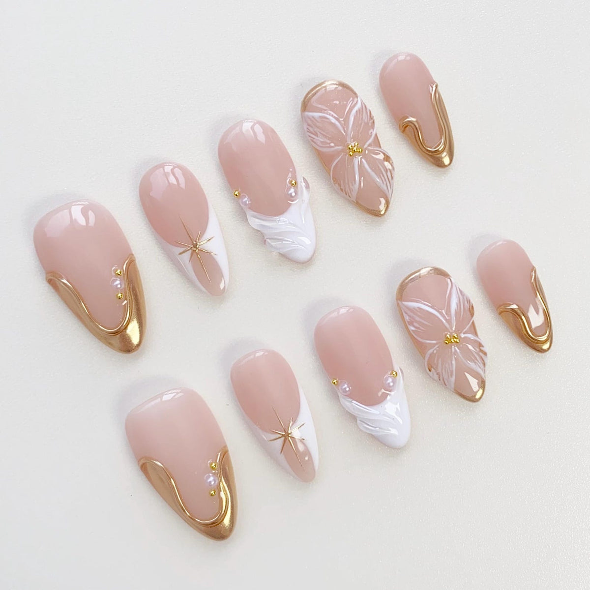 A set of almond-shaped artificial nails in various designs. The nails feature a pale pink base with gold accents, white floral and wave designs, and starburst details. Some nails have small pearl embellishments.