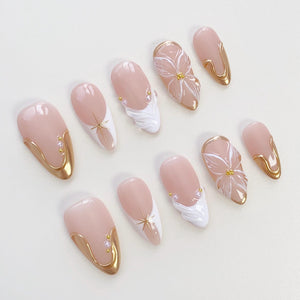 A set of almond-shaped artificial nails in various designs. The nails feature a pale pink base with gold accents, white floral and wave designs, and starburst details. Some nails have small pearl embellishments.