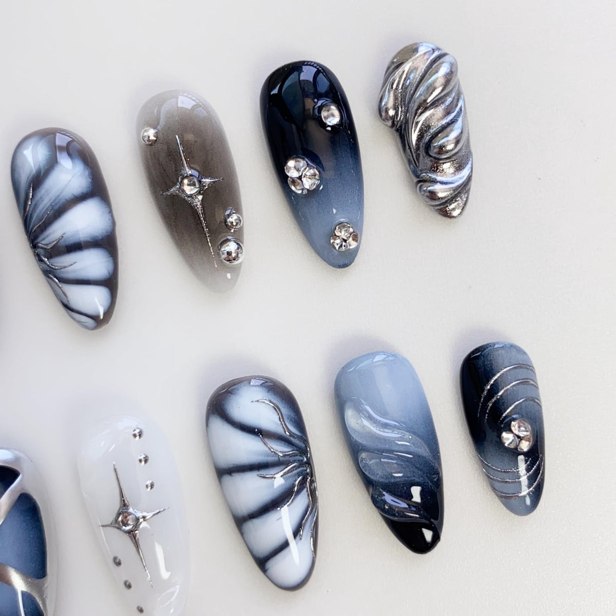 A set of ten artificial nails with various designs. The nails feature a mix of colors, including white, gray, and navy blue, with silver embellishments. Some nails have a swirl design, while others have star or floral patterns.
