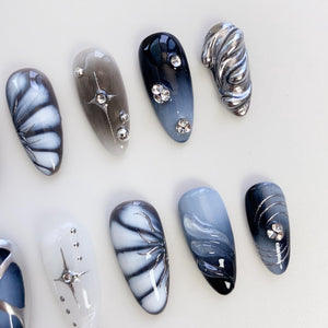 A set of ten artificial nails with various designs. The nails feature a mix of colors, including white, gray, and navy blue, with silver embellishments. Some nails have a swirl design, while others have star or floral patterns.