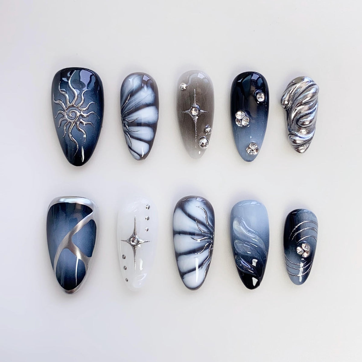 A set of ten artificial nails with various designs. The nails feature a range of colors, including dark blue, white, and silver. Some nails have intricate patterns, such as a sunburst, floral designs, and swirling effects. Embellishments include rhin