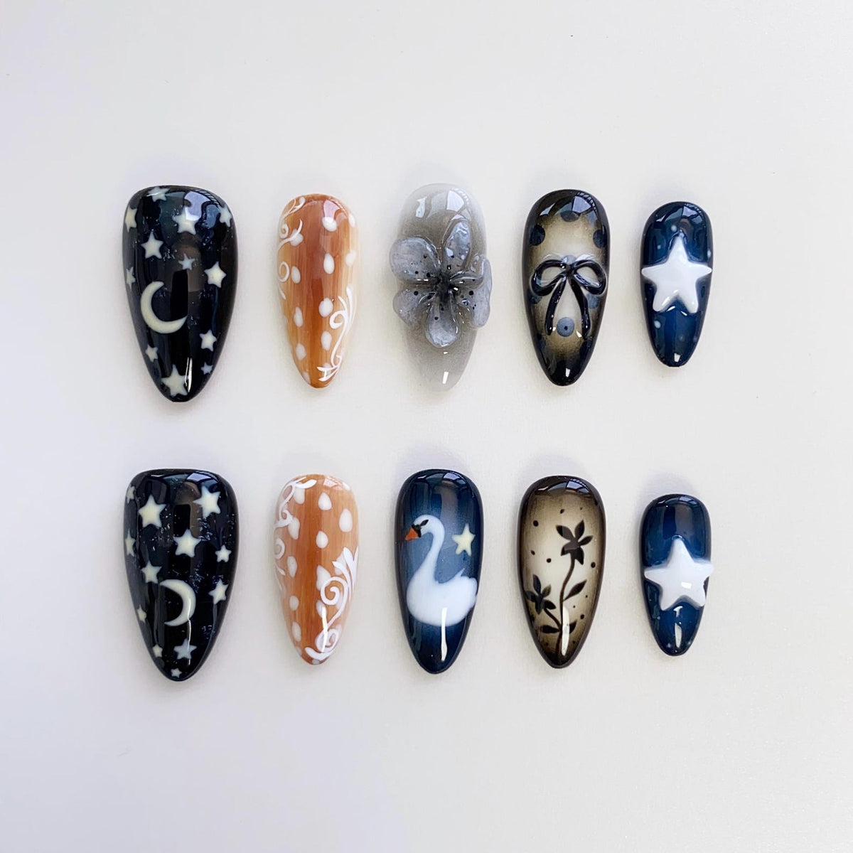 A set of ten artificial nails with various designs. The nails feature a dark blue base with white stars and a crescent moon, a brown base with white dots, and a clear base with a gray flower. Other nails have a swan, bow, and floral designs.