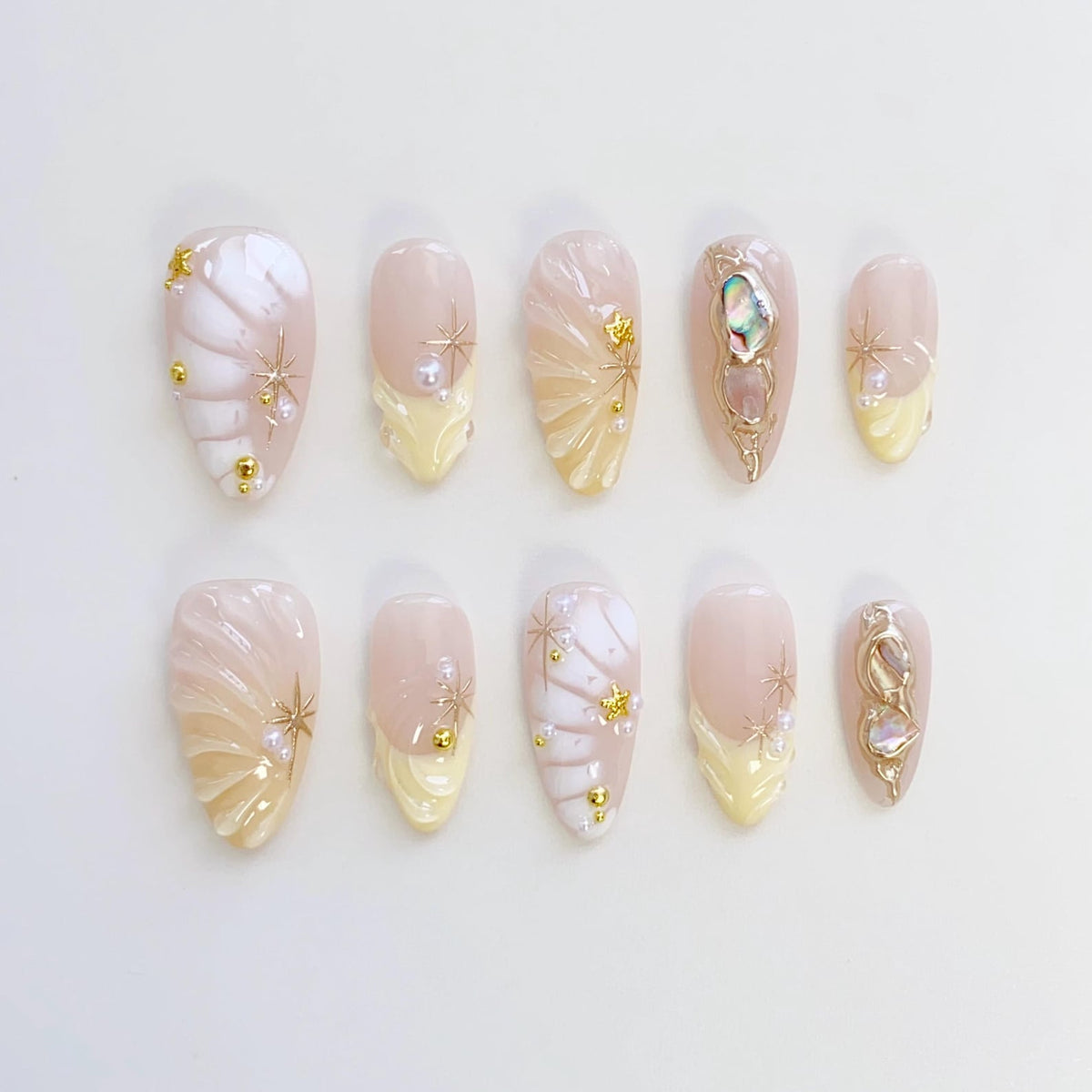 A set of ten almond-shaped false nails. The nails feature a light pink base with white and yellow accents. Some nails have gold star and pearl embellishments, while others have ornate gold designs with iridescent stones.