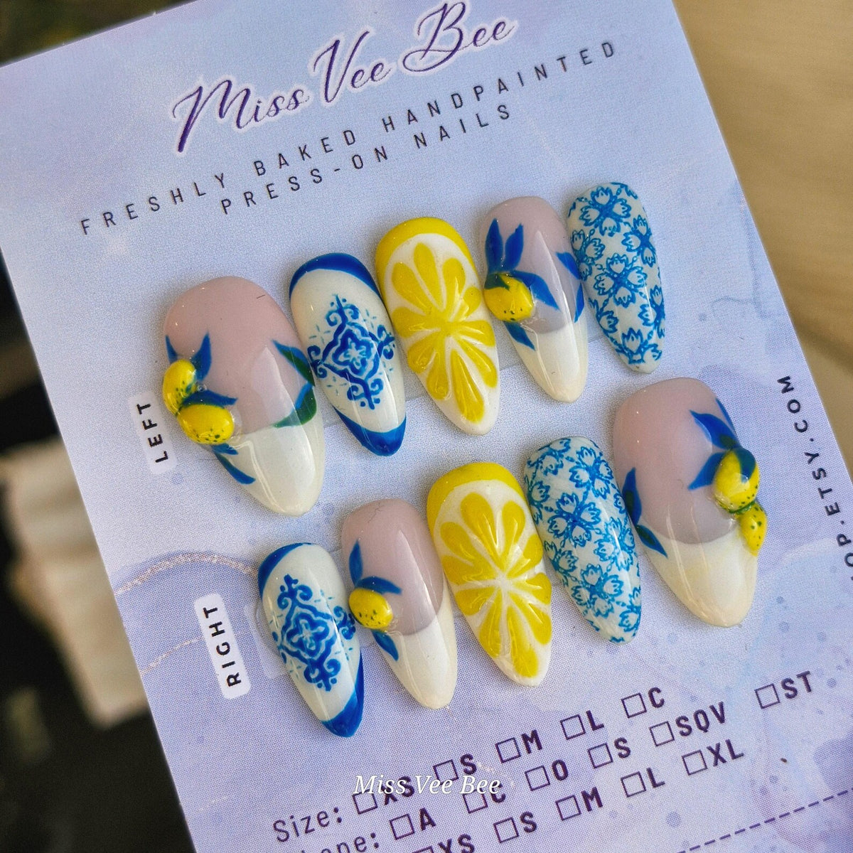 A set of hand-painted press-on nails with various designs. The nails feature a white base with yellow lemon slices, blue and white patterns, and lemon and leaf accents. The text "Miss Vee Bee" is visi