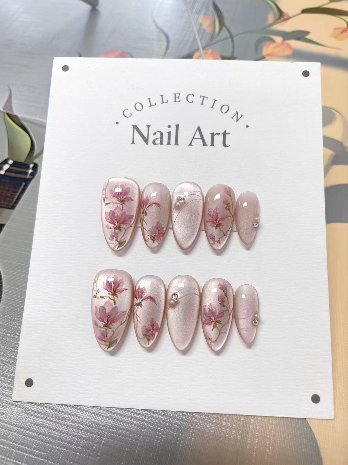 A set of ten almond-shaped press-on nails displayed on a white card labeled COLLECTION Nail Art. The nails feature a pearlescent light pink base with hand-painted pink floral designs, thin silver line
