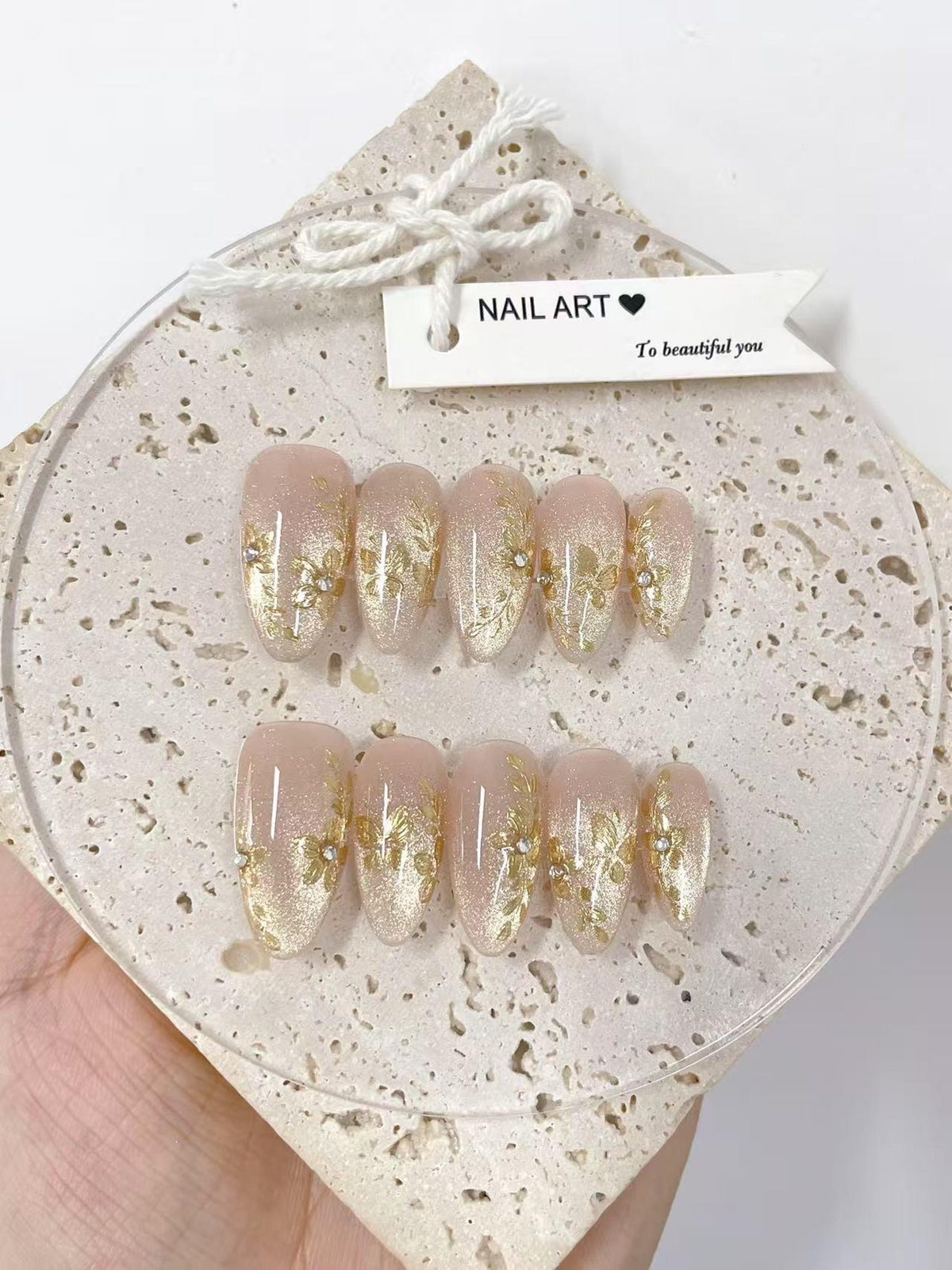 Set of oval-shaped false nails with a nude base and gold floral designs. Each nail is adorned with gold glitter and small rhinestones. The tag reads "NAIL ART" and "To beautiful you".