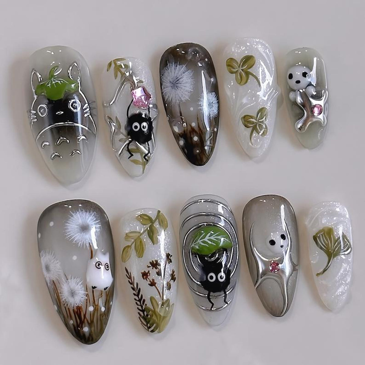 A set of ten artificial nails with various designs. The nails feature cartoon characters, dandelions, and floral patterns. Colors include white, gray, black, and green, with silver accents. The nails 