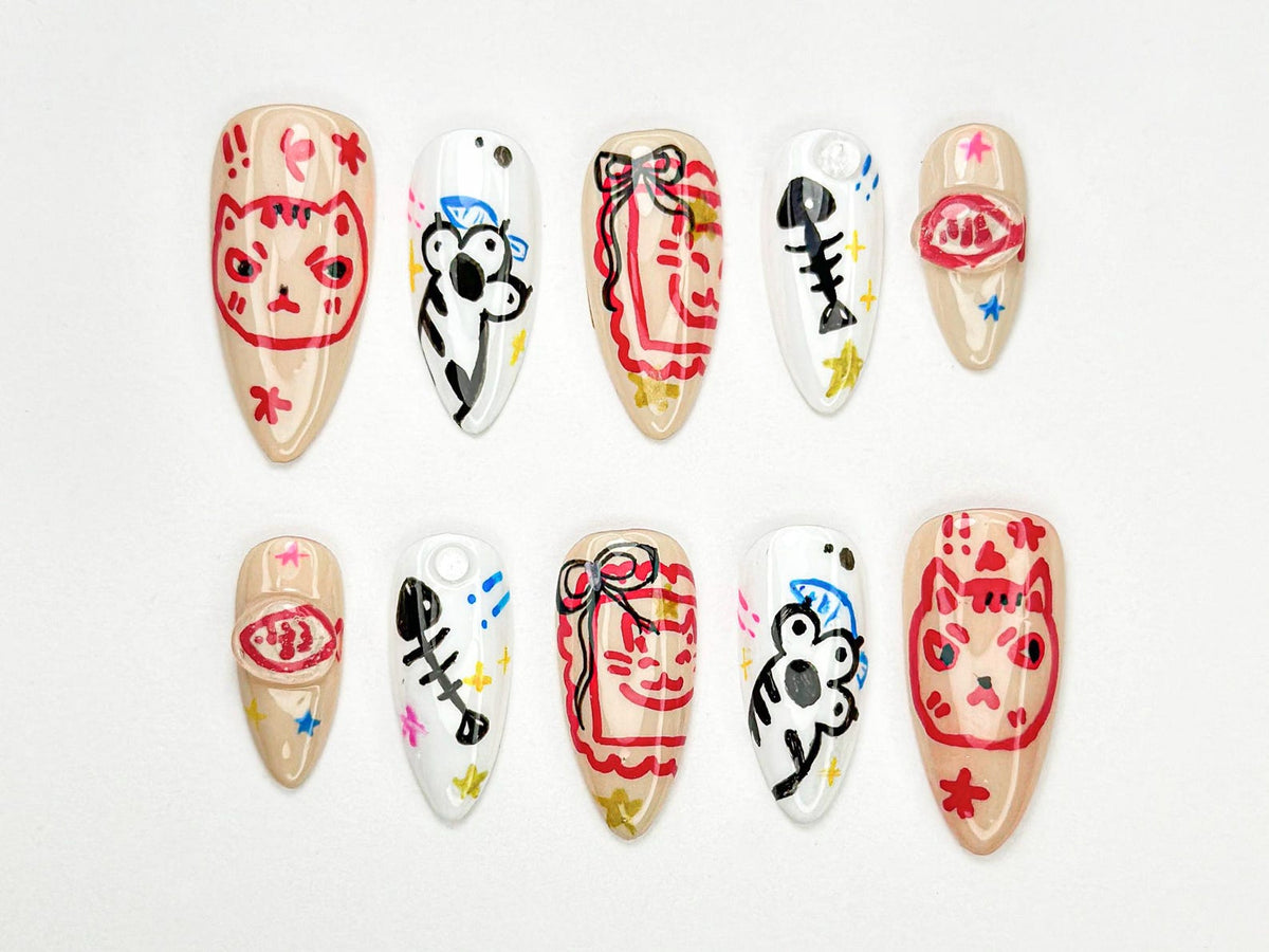 A set of ten almond-shaped artificial nails, each hand-painted with unique designs. The designs include a cat face, a koala, a fish skeleton, and abstract patterns in red, black, and blue on a neutral base.