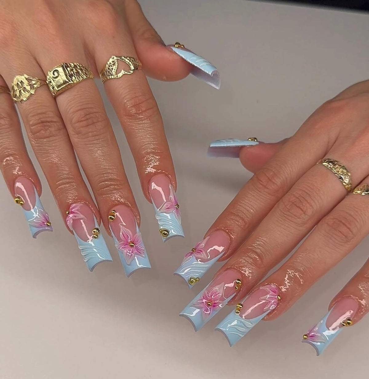 Long square-shaped artificial nails featuring a light blue French tip design with hand-painted pink floral accents and small gold metallic studs. The nails are worn with several gold rings on the fing