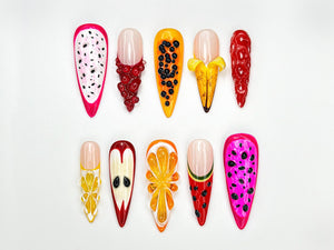 A collection of ten long, almond-shaped artificial nails, each uniquely designed with a fruit theme. The designs include dragon fruit, papaya, banana, and watermelon, featuring vibrant colors and intricate details. The nails are set against a white b