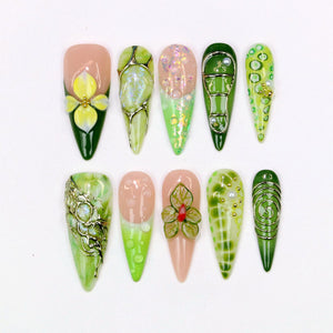 A collection of ten almond-shaped artificial nails with various designs. The nails feature a range of colors, including green, pink, and white, with floral and abstract patterns. Some nails have 3D embellishments and metallic accents.