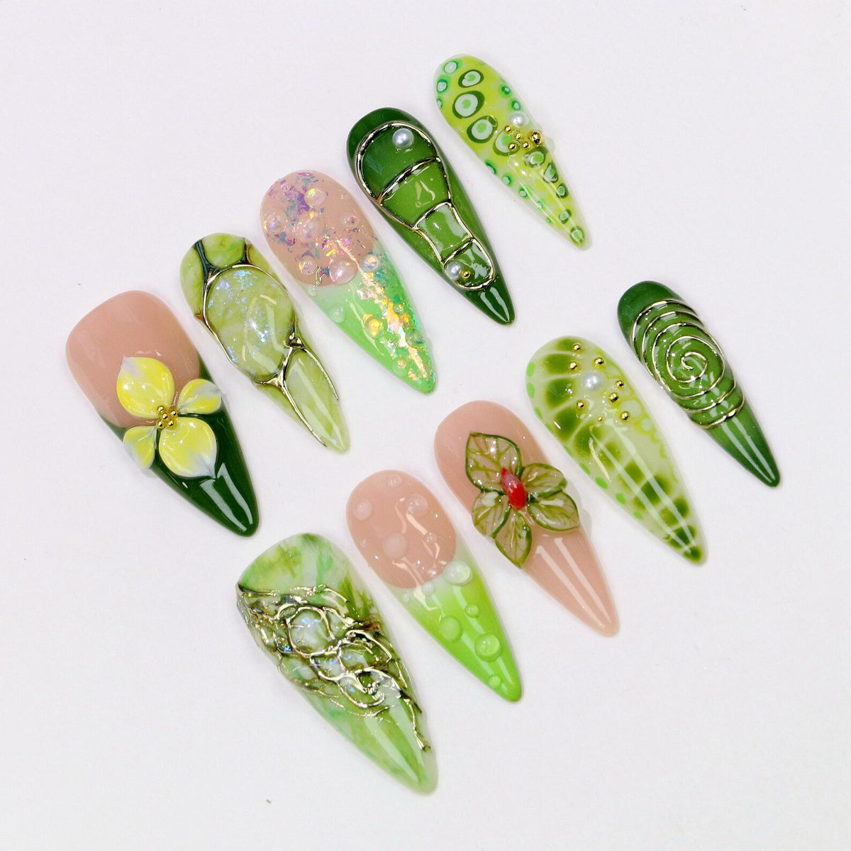 A collection of ten long, almond-shaped artificial nails. The nails feature various designs, including floral accents, glitter, and metallic wire embellishments. Colors include shades of green, pink, and gold, with a glossy finish.