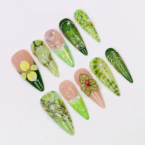A collection of ten long, almond-shaped artificial nails. The nails feature various designs, including floral accents, glitter, and metallic wire embellishments. Colors include shades of green, pink, and gold, with a glossy finish.