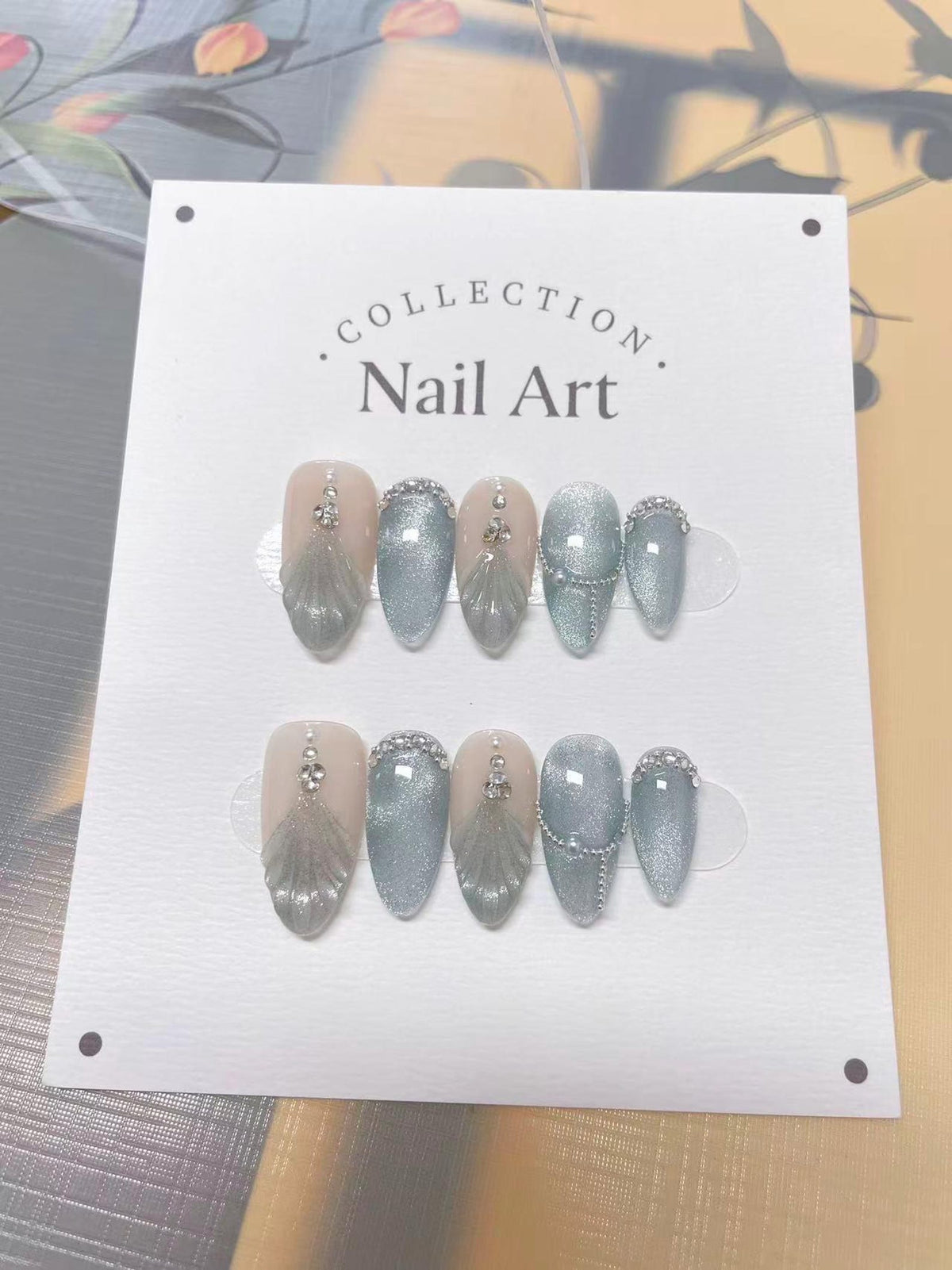 A set of artificial nails with a neutral base, adorned with silver glitter, rhinestones, and a feather design. The nails are oval-shaped and arranged on a white card with the text "Collection Nail Art