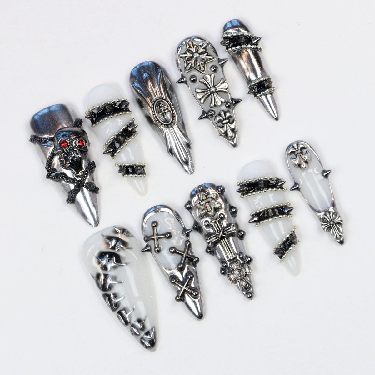 An assortment of silver-toned nail art, including skull, cross, and spike designs. The nail art pieces are displayed on a white surface, showcasing various gothic and edgy styles. The nails have a pointed shape.