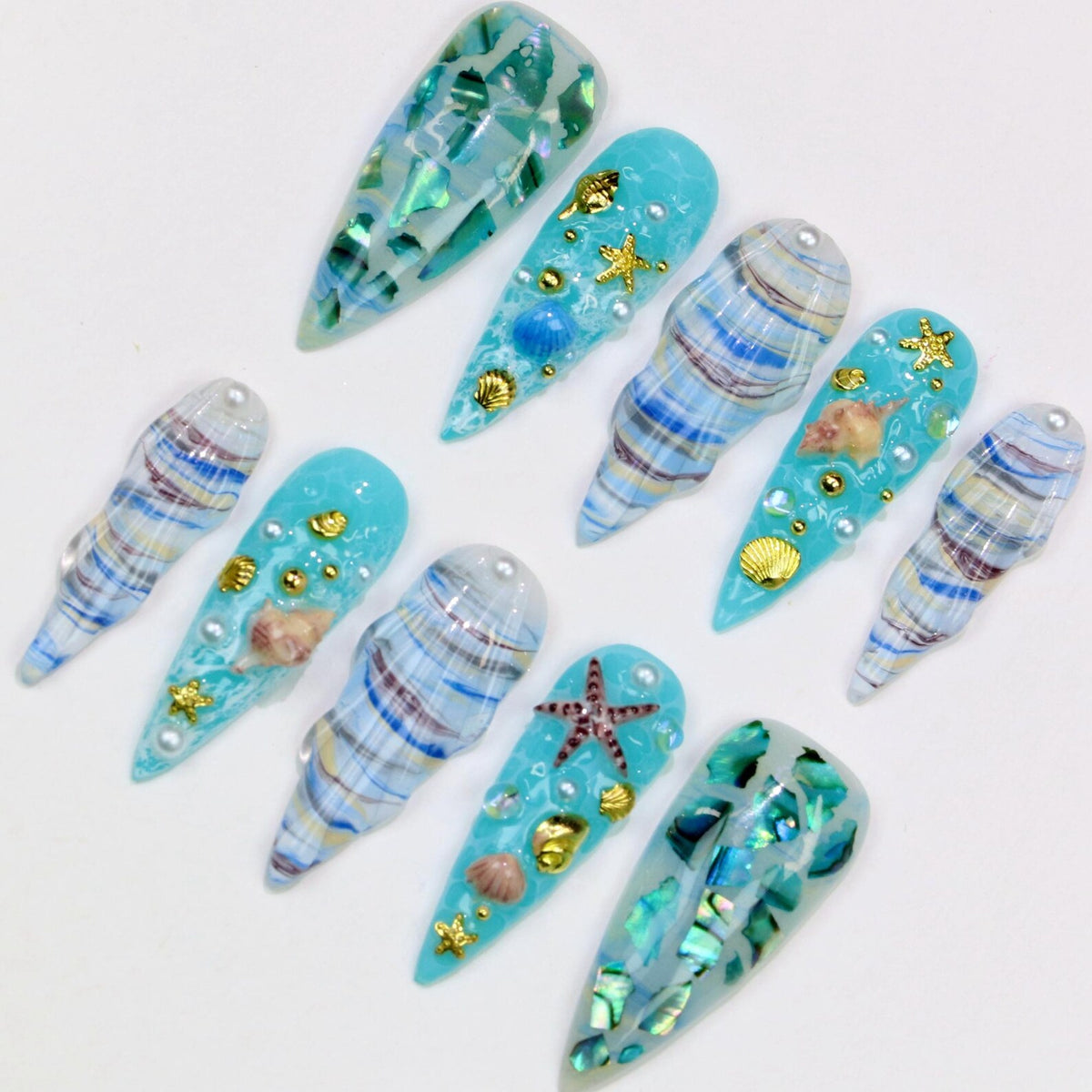 A collection of ten long, pointed artificial nails with a sea-themed design. The nails feature shades of blue, white, and gold, with seashell, starfish, and pearl accents. Some nails have a marbled effect, while others are adorned with iridescent pie