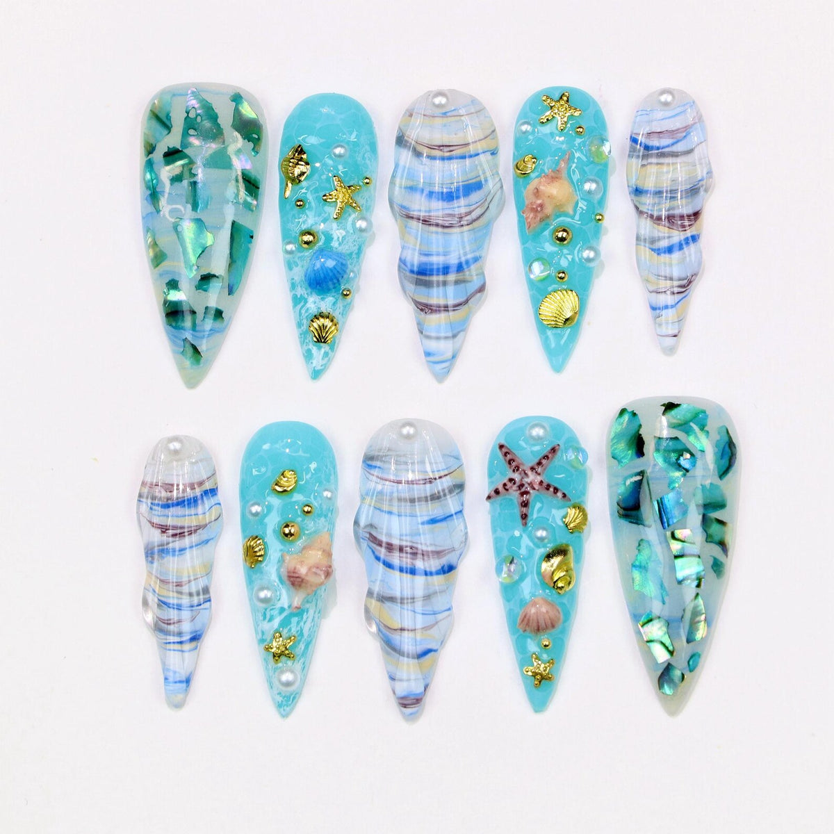 A collection of ten long, pointed artificial nails. The nails feature a variety of designs, including blue and white swirls, iridescent green and blue fragments, and seashell embellishments. Some nails have gold starfish and pearl accents.