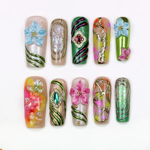 A collection of ten artificial nails with various designs. The nails feature floral and abstract patterns, with embellishments like glitter, gems, and 3D flowers. Colors include pink, blue, green, gold, and silver, creating a vibrant and artistic dis