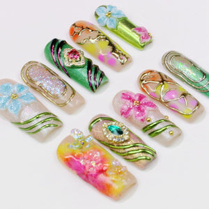 An assortment of artificial nails with various designs. Some nails feature floral embellishments in blue, pink, and yellow. Others have glitter, gems, and gold accents. The nails are in a variety of colors, including green, orange, and pink.