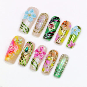 A collection of ten artificial nails, each with unique designs. The nails feature floral and abstract patterns, with accents of glitter, gold, and various colors including pink, green, blue, and yellow. Some nails have 3D floral embellishments.