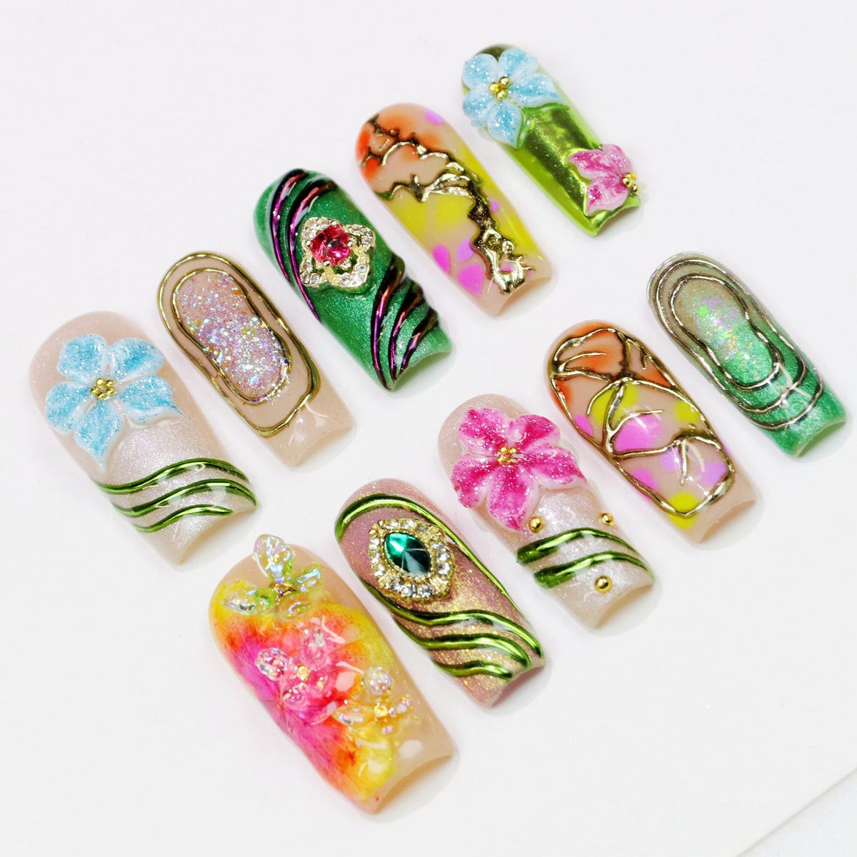 A collection of ten artificial nails, each uniquely designed with floral and jewel embellishments. The nails feature a variety of colors, including pink, green, blue, and gold, with intricate patterns and textures. The designs include glitter, gems, 