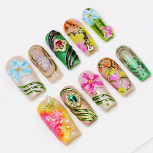 A collection of ten artificial nails, each uniquely designed with floral and jewel embellishments. The nails feature a variety of colors, including pink, green, blue, and gold, with intricate patterns and textures. The designs include glitter, gems, 