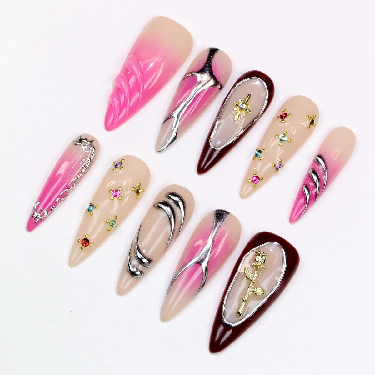A collection of ten long, almond-shaped artificial nails. The nails feature various designs, including pink gradients, silver accents, and jewel embellishments. Some nails have a textured appearance, while others showcase intricate metalwork and flor