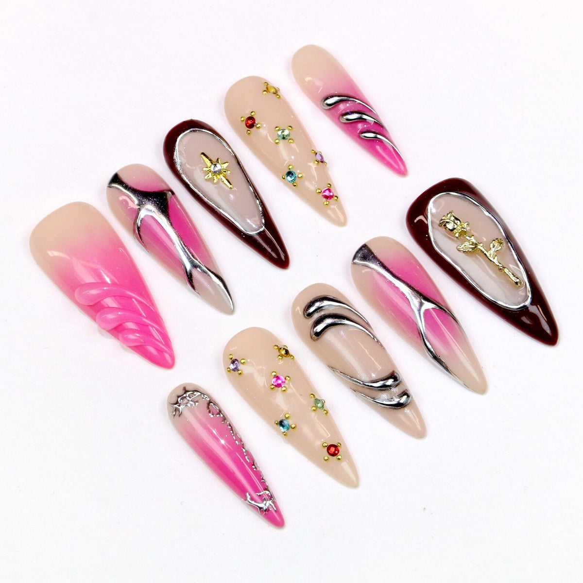 A collection of long, almond-shaped artificial nails. Designs include pink and nude gradients, silver accents, and jewel embellishments. Some nails feature a gold star or rose design. The nails are arranged on a white surface.