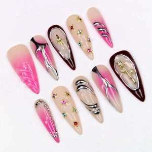 A collection of long, almond-shaped artificial nails. Designs include pink and nude gradients, silver metallic accents, and jewel embellishments. Some nails feature a gold rose or starburst design.