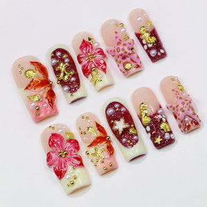 A set of ten artificial nails with various designs. The nails feature a light pink base with white tips and are adorned with 3D floral, starfish, and seashell embellishments in gold, red, and pink. Some nails have pearl accents.