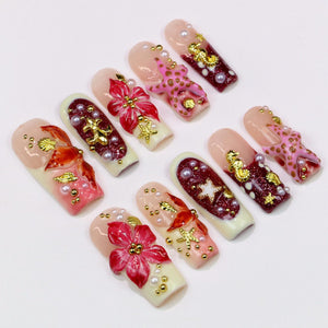 A collection of ten artificial nails with various designs. The nails feature a range of colors, including pink, red, and gold, with embellishments such as pearls, flowers, and starfish. The nails are arranged on a white surface.